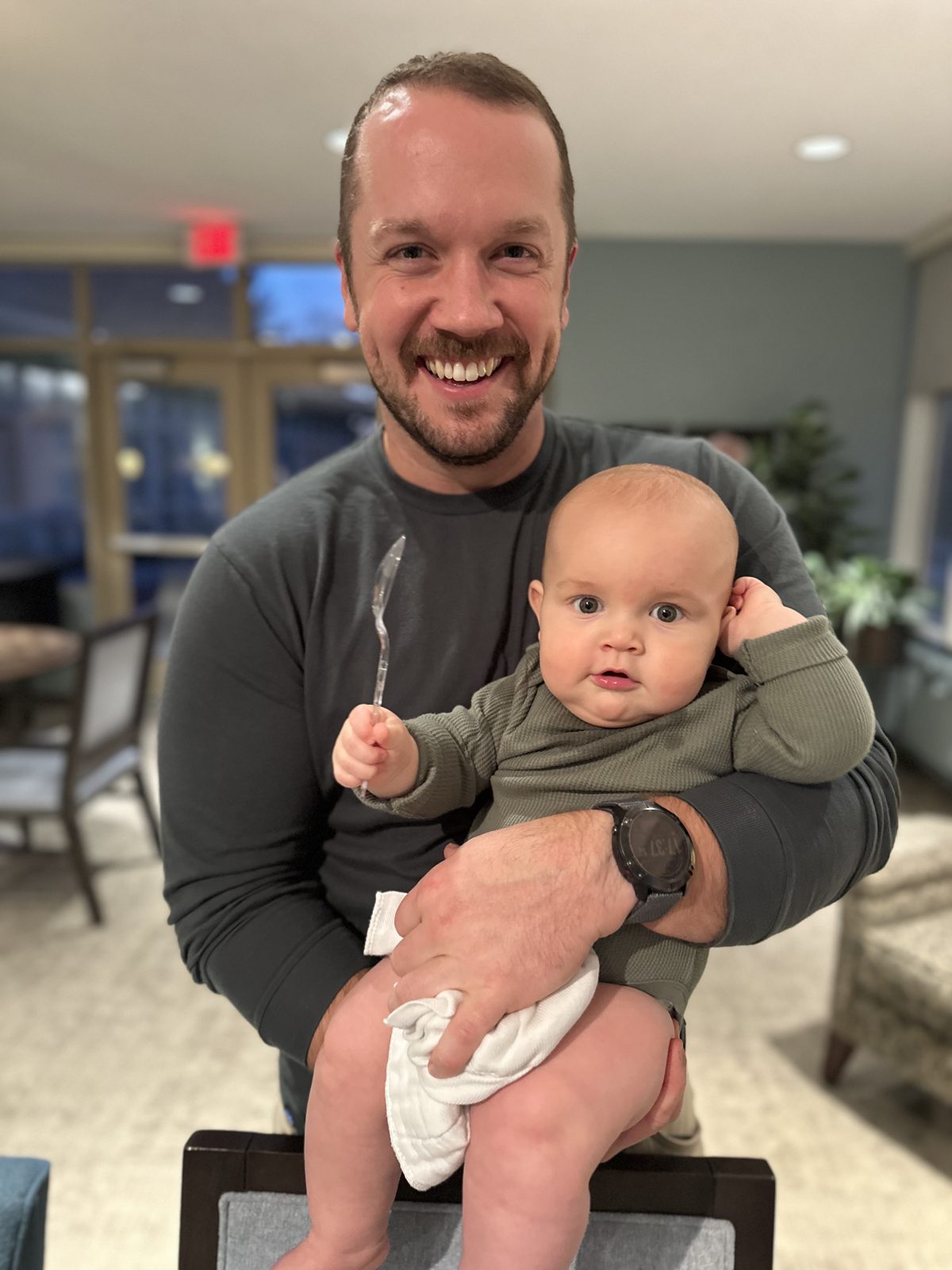 A smiling dad holding a baby who is gripping a spoon