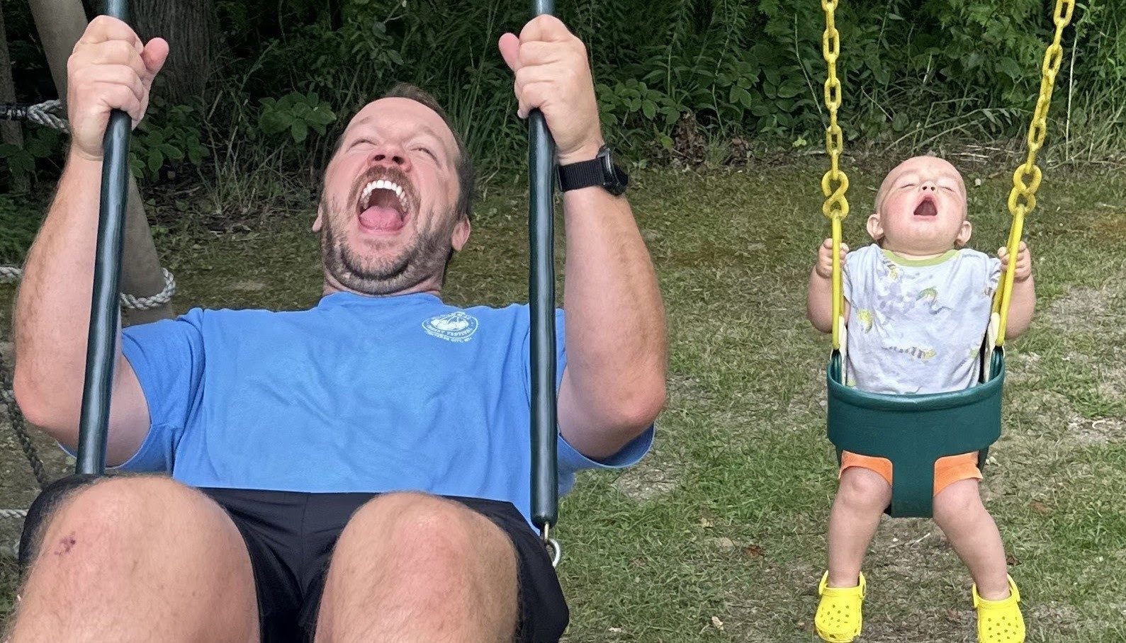 A man and a toddler laughing on side-by-side swings
